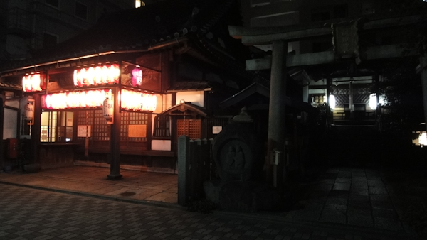 a building lit by many hanging laterns at night next to a stone torii gate